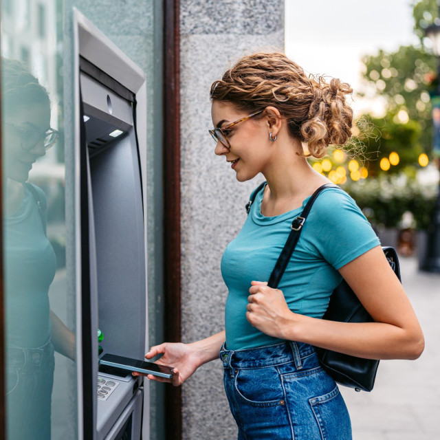 Young woman withdrawing money using phone at the ATM machine on the street in Budapest in Hungary.