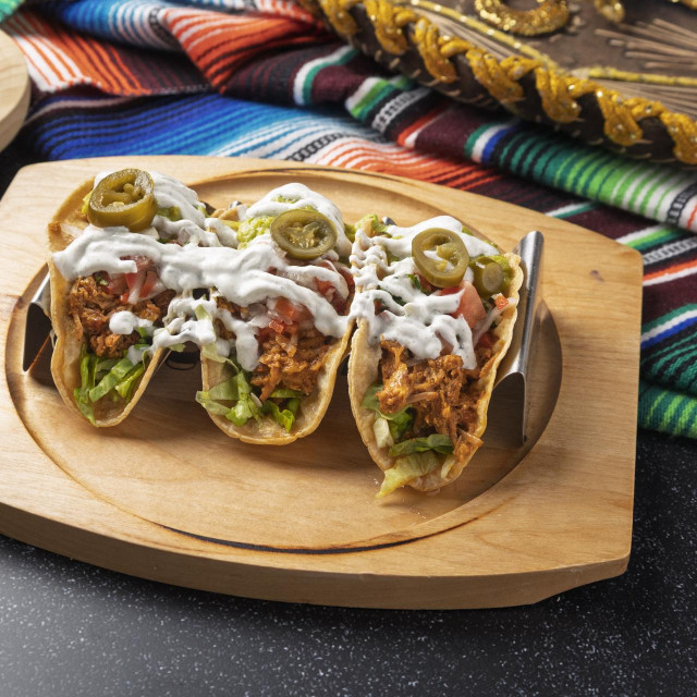 Three chicken tacos topped with jalapeños, lettuce, tomato, sour cream, and cheese are served on a wooden tray, accompanied by a traditional mexican sombrero and serape