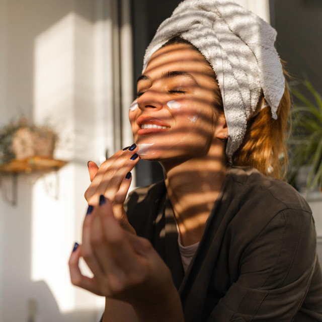 Young beautiful woman is sitting on the balcony nourishing her face with a hydrating cream. Woman has just washed her hair and has a towel on her head. Shadow striper are falling on beautiful woman. Woman selfcare at home.