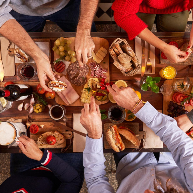Top view group of crop faceless friends eating delicious snacks while gathering at wooden table with beverages in light kitchen