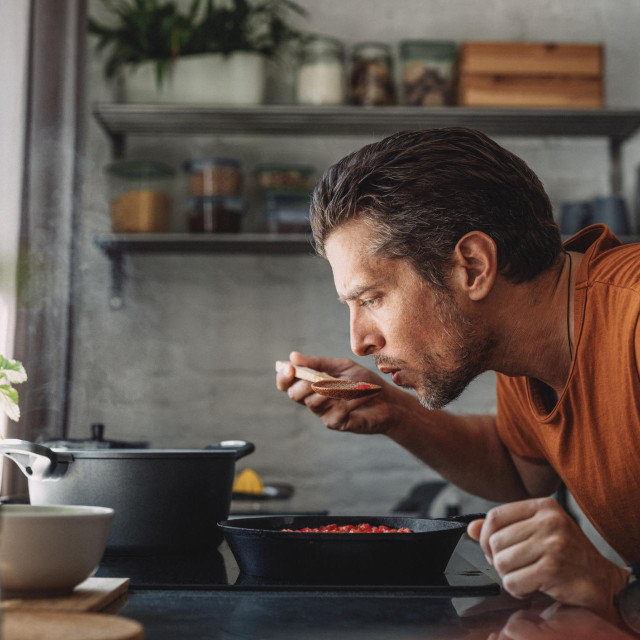 Close up shot of a handsome young happy Caucasian man tasting sauce with a mixing spoon with his eyes closed over a frying pan in a kitchen.