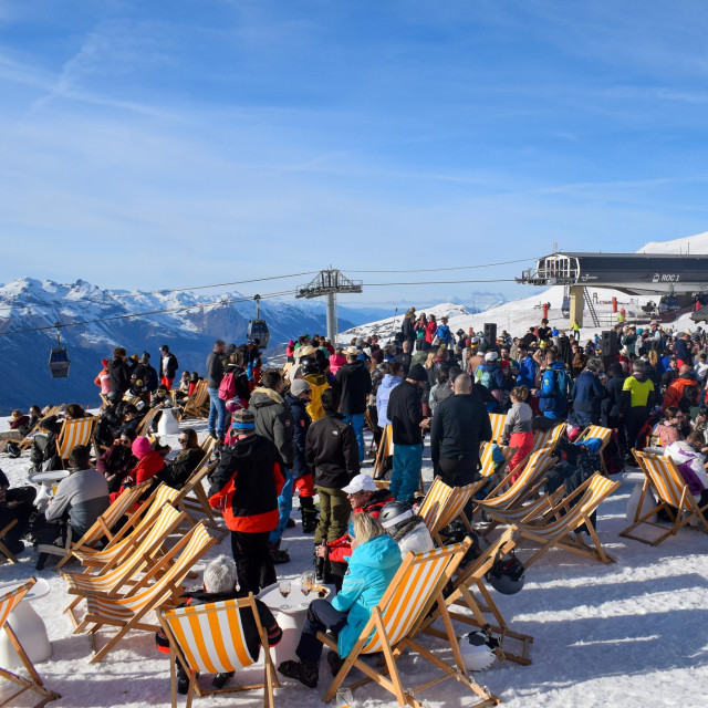 Les Menuires - March 13 2024: Lively apres ski party next to an active ski slope in Les Menuires French ski resort. Happy skiers drinking next to a breathtaking view of snowcapped Alpine mountains.