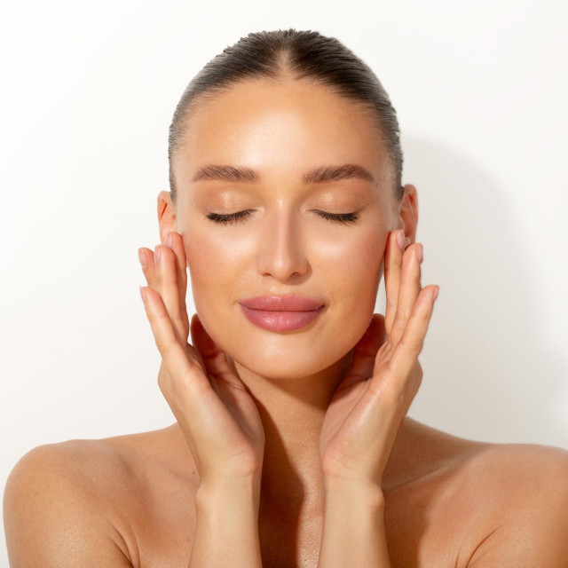 Perfect skin. Beautiful young European woman with nude makeup touching her cheeks with pleased face expression over white background