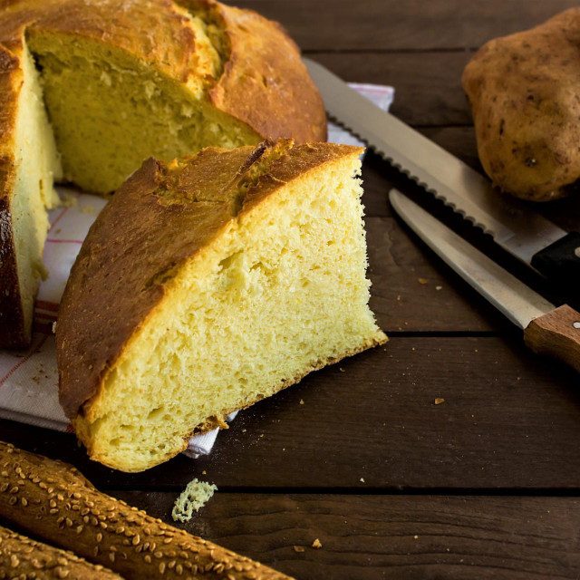 Above view of sliced potato bread on wooden table with knives and potatoes.