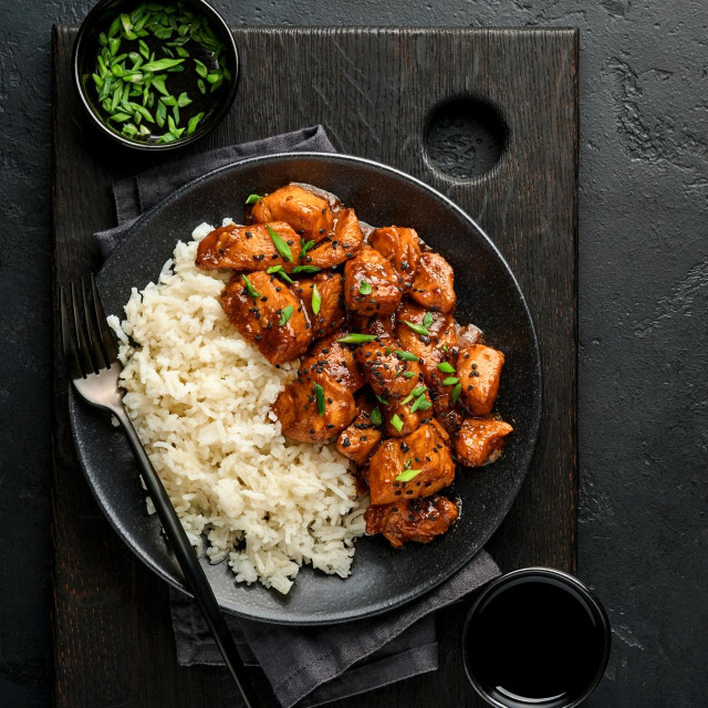 Spicy teriyaki chicken fillet pieces with rice, green onions and black sesame seeds on black plate on a dark slate, stone or concrete background. Top view with copy space.