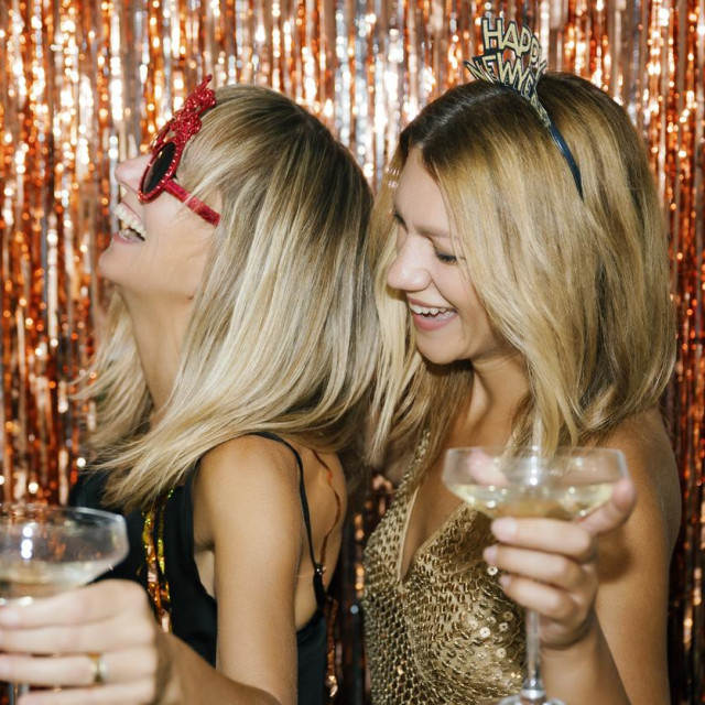 Two cheerful young women toasting with sparkling wine, wearing festive accessories and laughing in front of a shiny backdrop, joyfully celebrating new year‘s eve