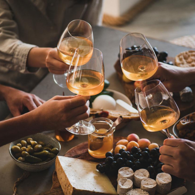 Glasses of white orange or rose wine in hands of celebrating friends over table with various gourmet snacks cheese grape bread. Gathering, celebrating, wine tasting and party concept