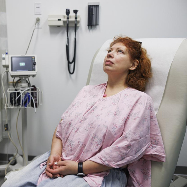 A 47-year-old Caucasian woman with reddish-brown hair sits on an examination table in a doctor‘s office. She wears a pink patterned hospital gown and has a thoughtful expression as she looks up. Her hands are clasped in her lap. The medical room includes a vital signs monitor and other diagnostic equipment in the background. This is a medium shot taken from an eye-level view, with available copy space to the left.