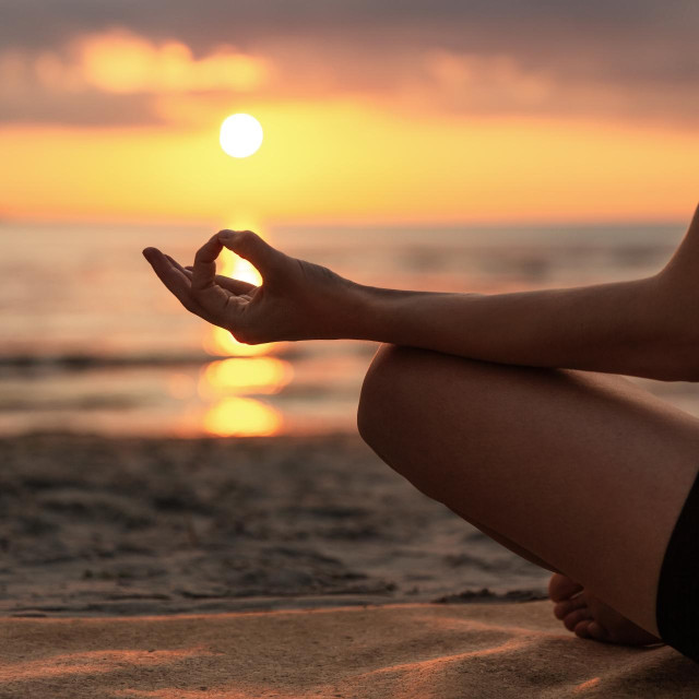 yoga, mindfulness and meditation concept - close up of woman meditating in lotus pose on beach over sunset