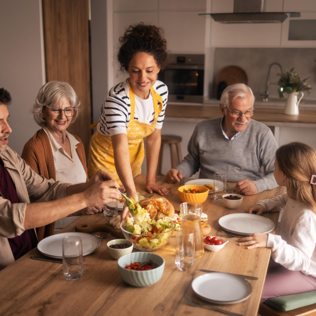 Multi-generational family gathered around a home dining table, adults and seniors sharing salad and roasted chicken, laughing and enjoying a casual lunch together