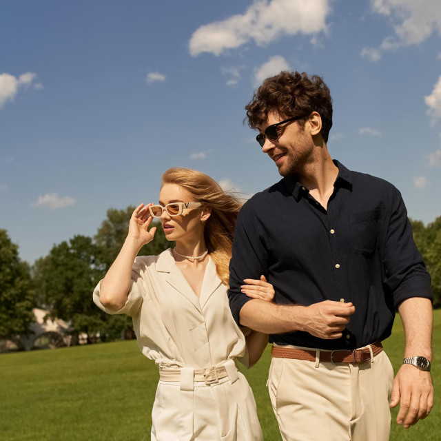A stylish duo, dressed in elegant attire, leisurely walk through a verdant field.