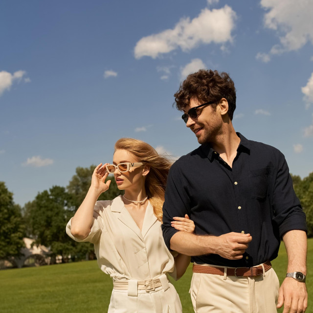 A stylish duo, dressed in elegant attire, leisurely walk through a verdant field.