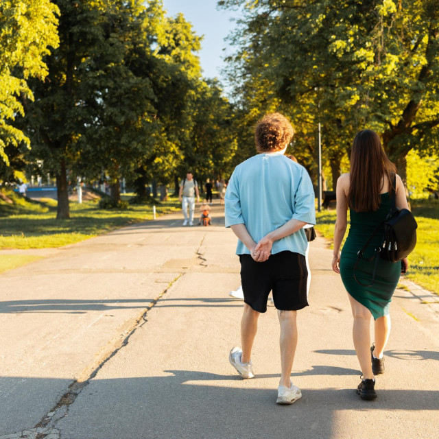 Young people stroll along a wide pathway in a city park surrounded by greenery. The sunlight enhances the tranquil atmosphere as they walk side by side.