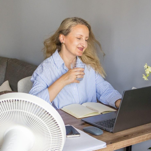 Woman enjoying cool airflow from a pedestal fan while using a laptop and holding a glass of water in a home office. Concept of summer comfort, healthy workplace, hydration and productive remote work.,Image: 1116982506, License: Royalty-free, Restrictions:, Model Release: yes, Credit line: Ina Yarashevich/Alamy/Profimedia