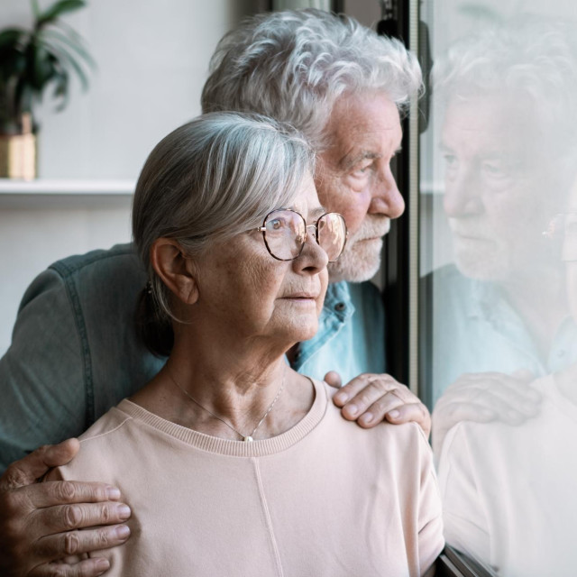 Pensive elderly mature senior man looking in distance out of window, thinking of personal problems. Old bearded male hugging sad wife close to him, copy space