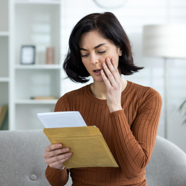Close-up photo of worried and shocked young woman sitting on sofa at home and reading received letter and message, holding hand to head in shock.