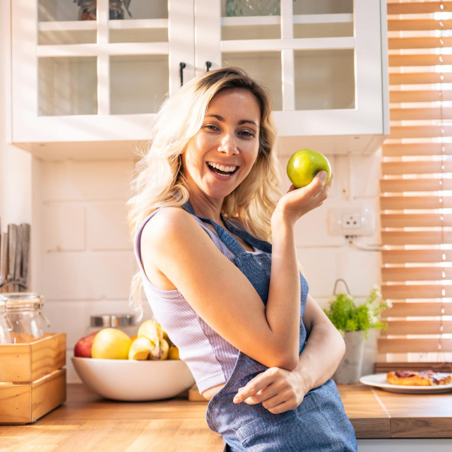 Caucasian young woman holding an apple on hands in the kitchen at home. Attractive beautiful girl wearing apron feeling happy and relax, enjoy cooking healthy fruit for lose weight and diet in house.