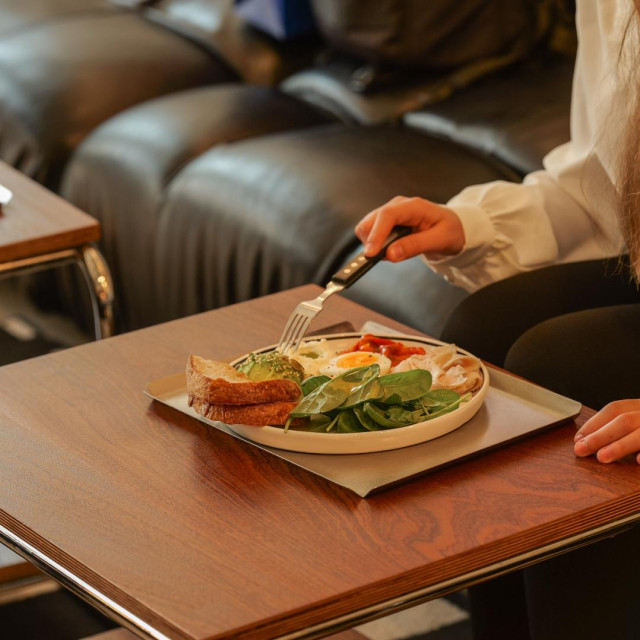 A person sits at a wooden table in a cafe, about to eat a nutritious meal consisting of eggs, avocado toast, and greens