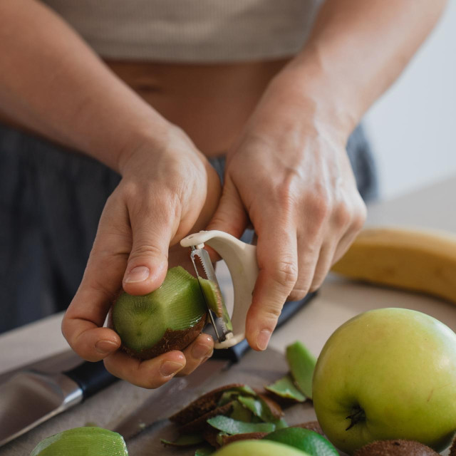 Woman Peeling Fresh Kiwi Fruit in Kitchen with Apples and Banana Nearby.