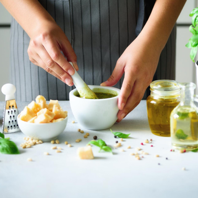 Woman hands making italian pesto in bowl. Ingredients - basil, lemon, parmesan, pine nuts, garlic, olive oil and salt on rustic wooden background. Top view, flat lay, copyspace