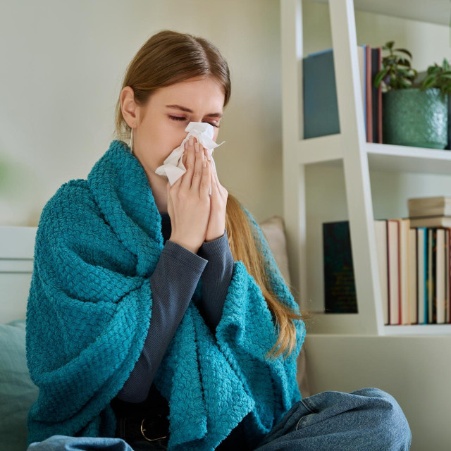 Young sick woman sitting under warm blanket sneezing coughing in handkerchief