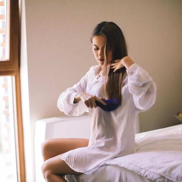 Young woman sitting on bed in bedroom and brushing her hair. She is beautiful and very sensual